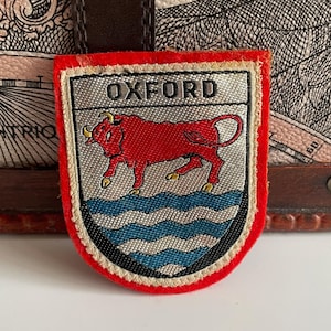 Könnte beinhalten: Schildförmiger Aufnäher mit dem Wort "OXFORD" oben. Der Aufnäher zeigt einen roten Bullen über blauen Wellenlinien, alles eingerahmt von einem silbernen Rand und einem roten Rand. Der Aufnäher ist wahrscheinlich ein Souvenir oder ein Emblem.