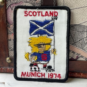 May include: A rectangular embroidered patch with a white background and black border. It features the red text SCOTLAND and MUNICH 1974. A cartoon character with yellow hair holds a blue and white Scottish flag above a soccer ball.