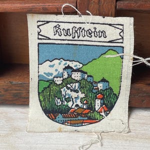 Könnte beinhalten: Ein Vintage-Stoffaufnäher mit dem Wort "Kufstein" oben. Der Aufnäher zeigt eine Landschaft mit Gebäuden, Bergen und einem grünen Hügel. Der Aufnäher hat einen weißen Rand und lose Fäden.