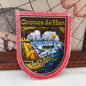 Puede incluir: Un parche rectangular con un borde rojo y una ilustración detallada de una escena de cueva. El parche presenta el texto "Grottes de Han" en letras amarillas. La imagen representa un río, botes y un acantilado amarillo y naranja.