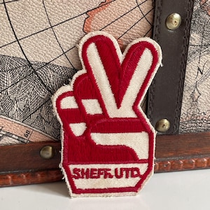 May include: An embroidered patch featuring a hand making a peace sign gesture in red and off-white thread. The design includes the text SHEFF UTD at the base of the hand. The patch has a thick red border and a flat, stitched texture throughout.
