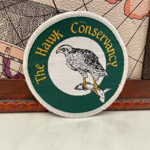 Könnte beinhalten: Runder Aufnäher mit grünem Rand und dem goldenen Schriftzug "The Hawk Conservancy". In der Mitte befindet sich eine detaillierte Illustration eines Falken mit einem Fisch in den Krallen, vor weißem Hintergrund.