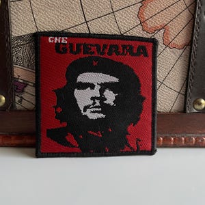 May include: A square patch with a red background and a black border. The patch features a black and white portrait of Che Guevara, with the name "CHE GUEVARA" in red letters at the top. It is likely an iron-on or sew-on patch.
