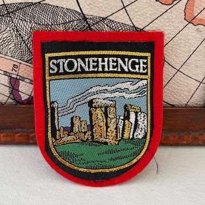 May include: Embroidered patch featuring Stonehenge. The patch has a red border, a black background, and a detailed image of the ancient monument. The word "STONEHENGE" is embroidered at the top. The patch is likely for a jacket or bag.