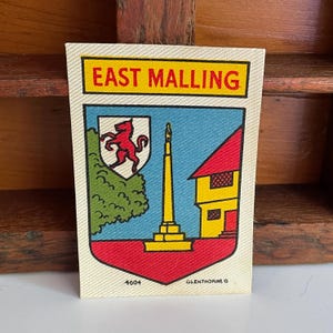 May include: A rectangular fabric patch featuring the text EAST MALLING in red on a yellow background. Below, a shield displays a red lion on a white field, a green tree, a yellow monument, and a yellow building on a red base, with 4604 and OLENTHORNE G.