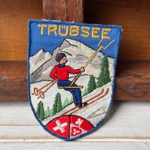 May include: Embroidered patch with the word "TRUBSEE" in yellow. The patch depicts a skier on a slope, mountains, and pine trees. The bottom of the patch has a red and white emblem.