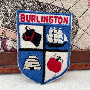 Puede incluir: Parche bordado con borde azul y la palabra "BURLINGTON" en letras rojas. El parche presenta cuatro cuadrantes: un toro negro, un barco, una colmena y una manzana roja. El parche es probablemente un recuerdo o un emblema.