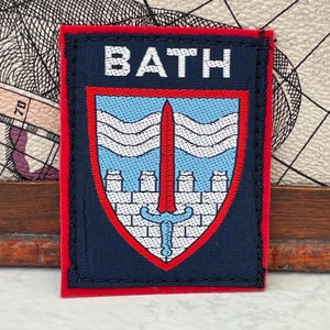 Könnte beinhalten: Ein rechteckiger gestickter Aufnäher mit dunkelblauem Hintergrund und rotem Rand. Das Wort BATH ist oben in Weiß gestickt. Darunter befindet sich ein Wappenschild mit hellblauem Feld, weißen Wellenlinien, einem Mauerwerk-Muster und einem roten Schwert.