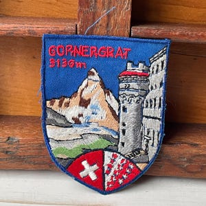 Può includere: Toppa ricamata con sfondo blu con la scritta "GORNERGRAT 3136m". La toppa raffigura una montagna, una torre e uno scudo con una croce e un motivo. La toppa è blu, rossa, bianca, verde e marrone.