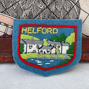 May include: A blue felt patch embroidered with the word HELFORD in yellow. The patch depicts a detailed scene of white buildings, green trees, and a red border.