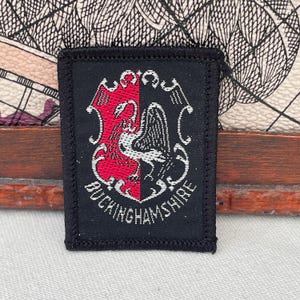 Könnte beinhalten: Rechteckiger schwarzer Aufnäher mit einem rot-weißen Emblem, das eine vogelartige Kreatur zeigt, und dem Text "BUCKINGHAMSHIRE" in Weiß. Der Aufnäher hat einen genähten Rand. Der Hintergrund ist gemustert.