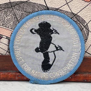 May include: A circular embroidered patch with a light blue border and a light gray center. The patch features a black silhouette of a lapwing bird and the text "LINCOLNSHIRE & SOUTH HUMBERSIDE TRUST FOR NATURE CONSERVATION" stitched in white.