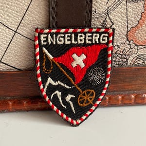 May include: Embroidered patch with the word "ENGELBERG" at the top. The patch features a red flag with a white cross, a snowflake, and a skier on a black background, framed by a red and white border. The patch is attached to a brown leather strap.