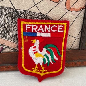 Puede incluir: Un parche rojo en forma de escudo con la palabra "FRANCE" en letras blancas. El parche presenta un gallo con detalles blancos, amarillos y rojos, y un diseño de rayas azules, blancas y rojas. El parche está delineado en amarillo.