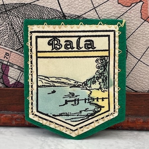 Può includere: Una toppa a forma di scudo con bordo in feltro verde e cuciture a zigzag gialle. Il centro presenta un pannello in tessuto color crema con la parola Bala stampata in nero. Un'illustrazione raffigura un lago, una riva e un piccolo molo.