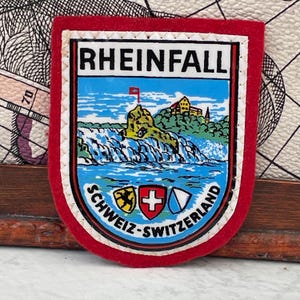 Puede incluir: Un parche de recuerdo en forma de escudo con un borde de fieltro rojo. El centro presenta una ilustración colorida de las cataratas del Rin, un castillo y tres pequeños escudos. El texto dice RHEINFALL y SCHWEIZ-SWITZERLAND en letras mayúsculas.
