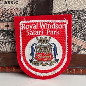 Könnte beinhalten: Ein rotes und weißes Royal Windsor Safari Park-Abzeichen. Das Abzeichen zeigt den Parknamen in weißer Schrift, eine Krone, zwei Delfine und ein detailliertes Emblem. Rechteckig mit abgerundeten Ecken und weißem Rand.