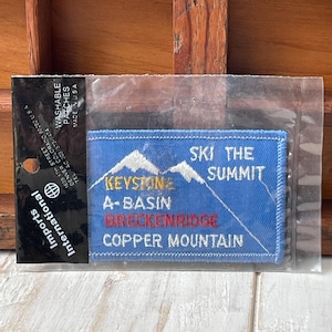 May include: A blue rectangular patch with white text and mountain graphics. The patch reads "SKI THE SUMMIT" and lists "KEYSTONE, A-BASIN, BRECKENRIDGE, COPPER MOUNTAIN". The patch is in a clear plastic sleeve.