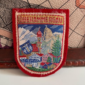 Puede incluir: Parche rectangular con borde rojo y escena bordada detallada. La escena presenta una iglesia, casas, montañas y el texto "OBERAMMERGAU". El parche es probablemente un recuerdo o un emblema.