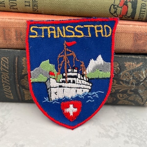 May include: A blue and red embroidered patch with the text "Strassstr" in yellow. The patch features a white ship sailing on blue water with green mountains in the background. A red shield with a white cross is in the lower center of the patch.