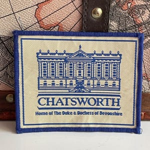 May include: A rectangular fabric patch with a cream-colored background and a blue embroidered border. The patch features a blue line-art illustration of Chatsworth House, with the text CHATSWORTH and Home of The Duke & Duchess of Devonshire.