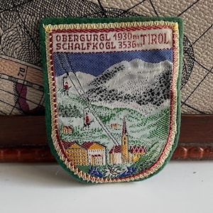 Könnte beinhalten: Vintage gestickter Aufnäher mit grünem Rand und Schildform. Der Aufnäher zeigt eine Berglandschaft mit Skilift, Gebäuden und dem Text "OBERGURGL 1930m TIROL SCHALFKOGL 3536m". Der Aufnäher hat ein Farbschema aus Rot, Gelb, Blau und Weiß.