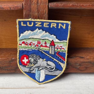 May include: Embroidered patch with the word LUZERN at the top. The patch features a blue background with a mountain range, a lake, and a lion. The patch has a red and white Swiss flag and a shield with blue and white stripes.