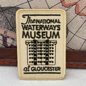 May include: A rectangular patch with the text "The National Waterways Museum at Gloucester" in black on a cream background. The patch features an illustration of a building. The patch has a stitched border.