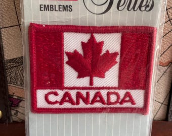 May include: A red and white embroidered patch featuring a red maple leaf and the word "CANADA" in red lettering. The patch is on a white background with the words "CANADA EMBLEMS" printed above it.