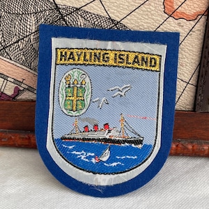 Op de afbeelding: Een blauwe patch met de woorden "HAYLING ISLAND" in gouden letters. De patch toont een afbeelding van een schip op blauw water, zeemeeuwen en een wapen. De patch heeft een witte rand.