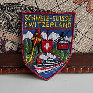 May include: Embroidered patch with the text "SCHWEIZ-SUISSE SWITZERLAND" in yellow on a blue and red shield. The patch features images of a cable car, a boat, and a Swiss flag.