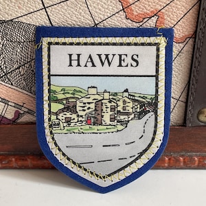 May include: A shield-shaped fabric patch with a blue border and yellow stitching. The center features a printed illustration of a town scene with stone buildings, green hills, and a road, with the word HAWES printed in black capital letters above.