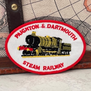 Puede incluir: Parche ovalado con borde rojo y fondo blanco. El parche presenta una ilustración de tren de vapor en negro y verde oliva, con el texto "PAIGNTON & DARTMOUTH" arriba y "STEAM RAILWAY" abajo, todo en rojo.