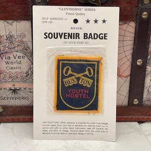 May include: A woven souvenir badge from the "Glenthorne" series. The badge is rectangular with a navy blue background and a gold border. It features the text "BUXTON YOUTH HOSTEL" and two crossed keys. The badge is self-adhesive or sew-on.