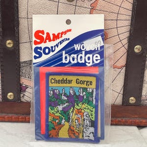 May include: A souvenir badge from Sampson Souvenirs featuring a colorful image of Cheddar Gorge. The badge is woven and comes in a clear plastic package with a red backing. The packaging also includes the text "woven badge".