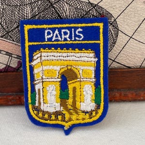 May include: Embroidered patch featuring the Arc de Triomphe in Paris, France. The patch is blue with a yellow border and the word "PARIS" in white. The monument is depicted in yellow, brown, and white.