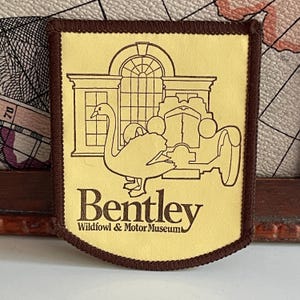 May include: A rectangular patch with a light yellow background and a dark brown border. The design features a line drawing of a swan in front of a building and a vintage car. The text reads Bentley Wildfowl & Motor Museum in dark brown.