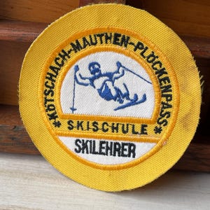 Puede incluir: Un parche redondo amarillo con un centro blanco que presenta un gráfico de un esquiador azul. El parche tiene texto en un patrón circular que dice "KOTSCHACH-MAUTHEN-PLOCKENPASS" y "SKISCHULE SKILEHRER".