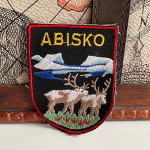 Peut inclure: Écusson brodé avec le mot "ABISKO" en lettres dorées. L'écusson représente un paysage de montagne avec des rennes et une bordure rouge. Le fond est noir.