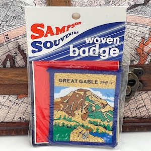 May include: A blue and gold woven badge with the text "Great Gable 2949 ft" and a depiction of a mountain. The badge is packaged in a clear plastic bag with a red and blue header that reads "Sampson Souvenir woven badge".