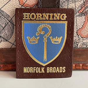 Peut inclure: Une étiquette de bagage rectangulaire en cuir marron avec un écusson bleu orné d'une crosse dorée et de deux couronnes dorées. Le texte HORNING est imprimé en doré au-dessus de l'écusson et NORFOLK BROADS en dessous. L'étiquette a des bords cousus.