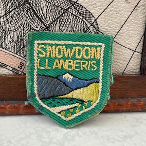 May include: A shield-shaped embroidered patch with a green background featuring the yellow stitched text SNOWDON LLANBERIS. The design includes a mountain landscape with blue and grey peaks, a white snow-capped summit, and a yellow grassy foreground.