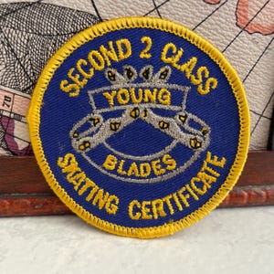 Pode incluir: Um patch azul circular com texto e borda bordados em amarelo. O texto diz "SECOND 2 CLASS", "YOUNG BLADES" e "SKATING CERTIFICATE". O patch apresenta um desenho de dois patins.