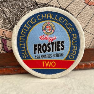 May include: A round patch with a blue border and the words "SWIMMING CHALLENGE AWARD" in yellow. The center features the text "Kellogg's FROSTIES" and "ASA AWARDS SCHEME" with a red stripe at the bottom that says "TWO".