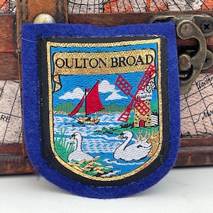Könnte beinhalten: Ein blauer Filz-Patch mit goldener Stickerei, der eine Szene aus Oulton Broad darstellt. Die Szene zeigt eine Windmühle, ein Segelboot, Schwäne und Wasser. Der Text "Oulton Broad" ist in Gold gestickt.