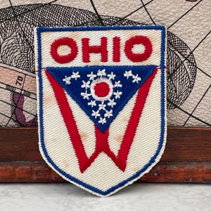 以下が含まれることがあります： 盾の形をした刺繍ワッペンで、上部に赤い文字でOHIOと書かれています。その下には青い三角形のフィールドがあり、中央の赤い円を17個の白い星が囲んでいます。クリーム色の背景に大きな赤いV字型が描かれています。