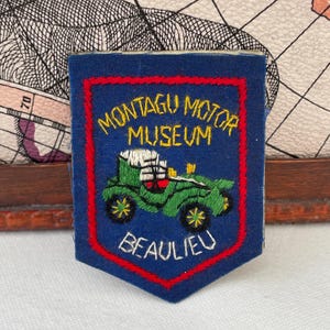 May include: A blue embroidered patch with the text "Montagu Motor Museum Beaulieu". The patch features a green vintage car design with yellow and red accents, framed by a red border.
