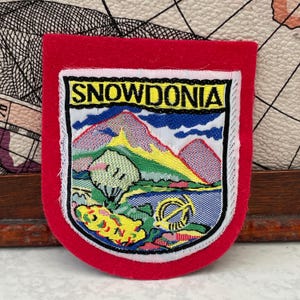 Puede incluir: Un parche bordado en forma de escudo con un borde rojo brillante. El centro presenta un paisaje con montañas, un lago y vegetación. La palabra SNOWDONIA está bordada en letras negras sobre un fondo amarillo en la parte superior del parche.