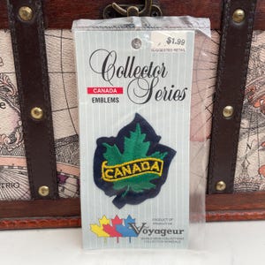 May include: A "Collector Series" emblem with a green maple leaf and yellow "CANADA" lettering on a dark blue background. The emblem is in a clear plastic packet. The text "CANADA EMBLEMS" is also visible.