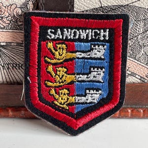 May include: Embroidered patch with a shield design. The patch has a red border, black backing, and the word "SANDWICH" at the top. The shield is divided into red and blue sections, with gold and white emblems.